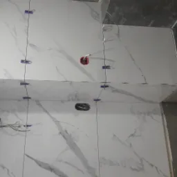 Białe płytki imitujące marmur, ułożone na ścianach i suficie łazienki, z widocznymi niebieskimi krzyżykami dystansowymi, otworami na gniazdka elektryczne i wystającymi przewodami.