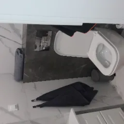 Nowoczesna toaleta podwieszana z szarą spłuczką na tle marmurowych płytek, uzupełniona o szare ręczniki i dywanik.