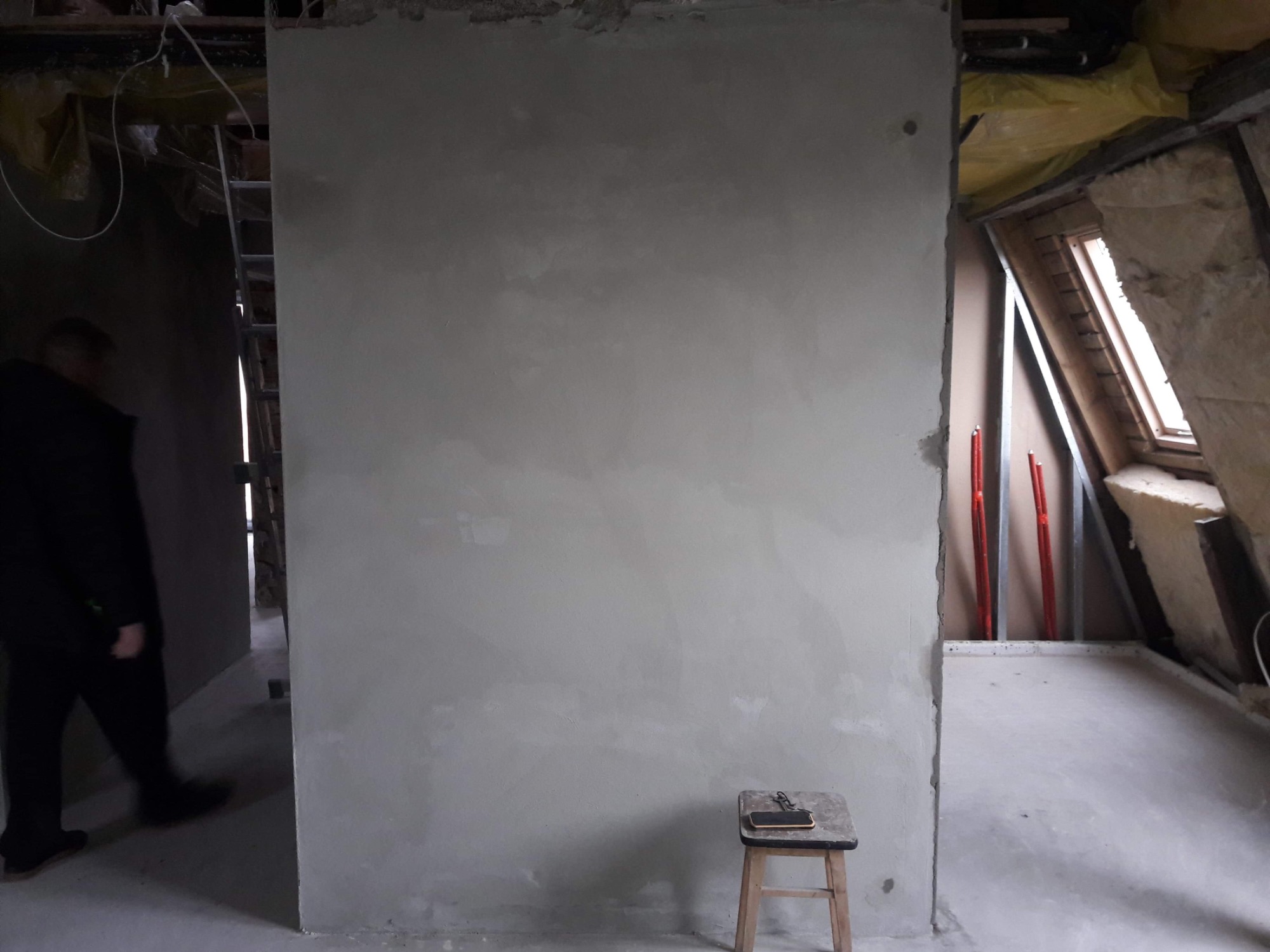 Świeżo otynkowana ściana działowa w surowym wnętrzu poddasza, z widocznym oknem dachowym i izolacją, na stołku leży telefon komórkowy i klucze.