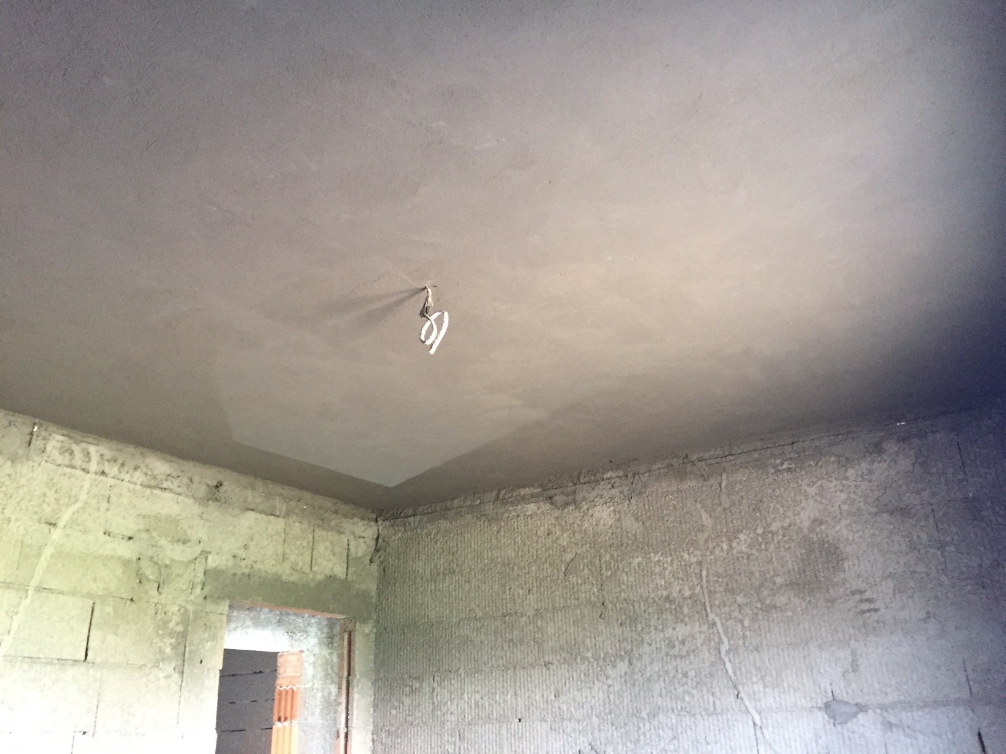 Surowy stan wnętrza z szarymi ścianami z bloków i świeżo otynkowanym sufitem z wystającym kablem elektrycznym pośrodku.