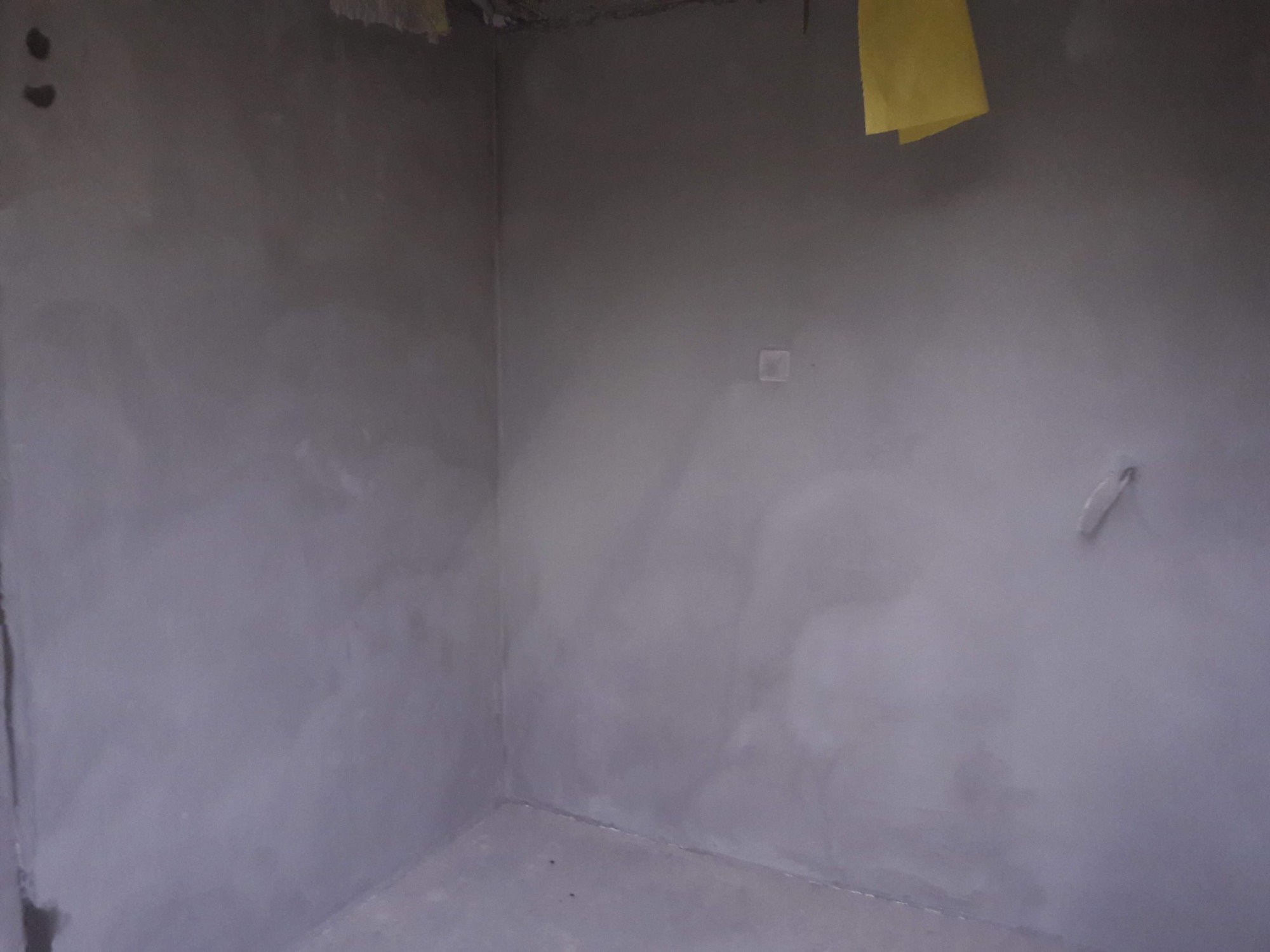 Świeżo położone szare tynki maszynowe w rogu pomieszczenia, widoczne ślady po szpachlowaniu, żółta taśma malarska przy suficie, mała puszka elektryczna na ścianie.