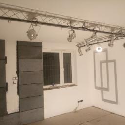 Zip-Bud - Montaż szarych, prostokątnych płytek na ścianie w trakcie prac wykończeniowych, widoczne oświetlenie sufitowe i szare kontury dekoracyjne na ścianie.