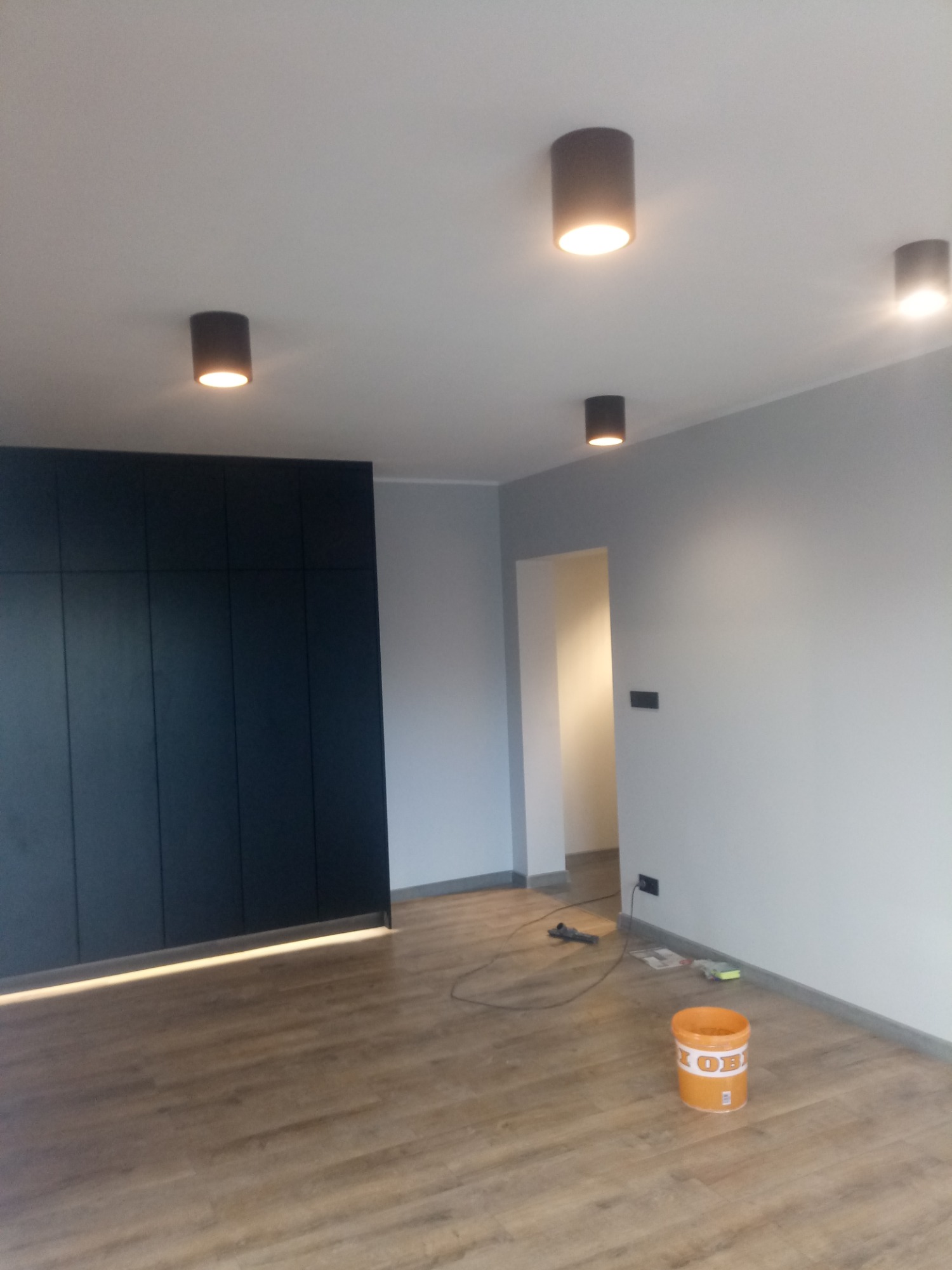 Pomieszczenie po malowaniu ścian na szaro, z zamontowanym oświetleniem punktowym i szafą w zabudowie, widoczne wiadro z farbą na podłodze.
