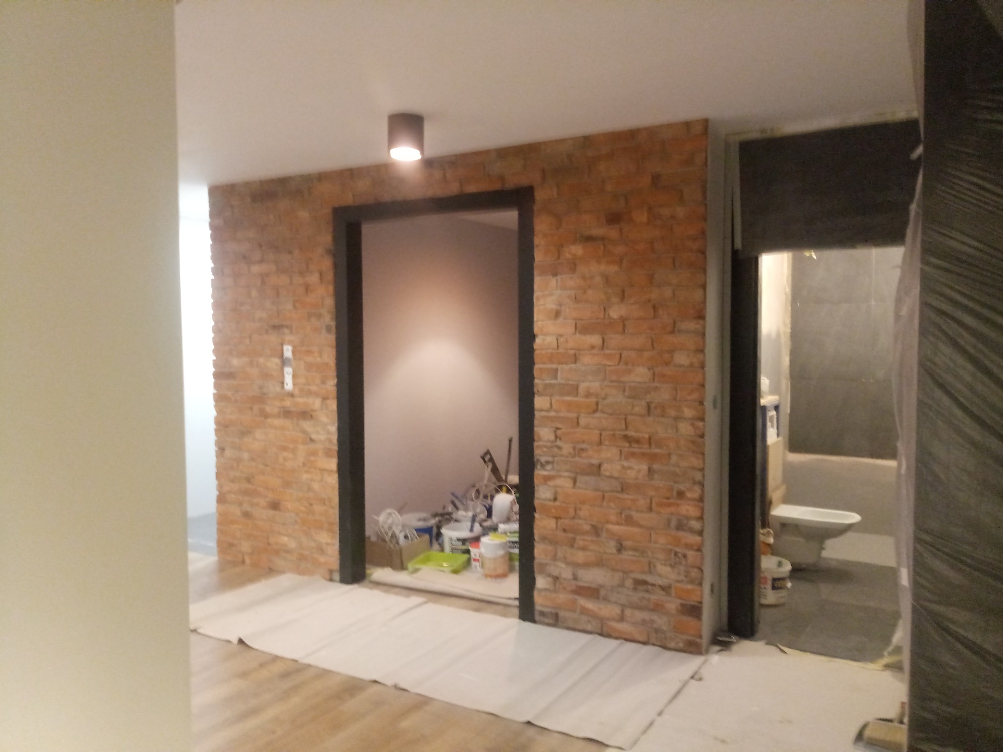 Ceglana ściana z czarną framugą drzwi, widok na remontowane wnętrze z narzędziami i pojemnikami, podłoga zabezpieczona folią, w tle widoczna toaleta w trakcie remontu.