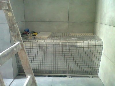 Niedokończona zabudowa z mozaiki w łazience, widoczna drabina i narzędzia glazurnicze, szare płytki ścienne w tle.