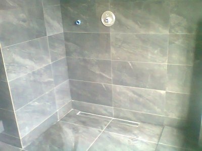 Wykończony brodzik z szarymi, marmurkowymi płytkami, odpływ liniowy w podłodze, widoczne przyłącza wodne na ścianie.