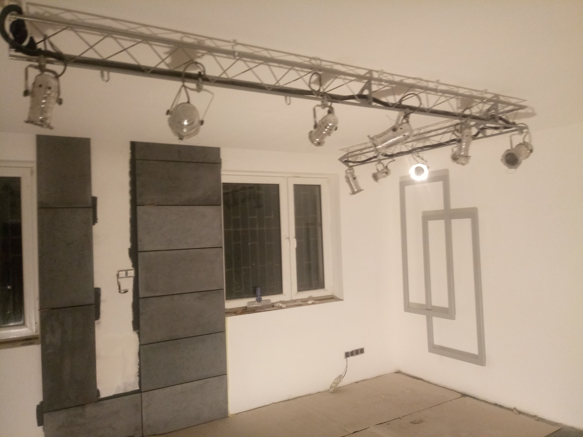 Montaż szarych, prostokątnych płytek na ścianie w trakcie prac wykończeniowych, widoczne oświetlenie sufitowe i szare kontury dekoracyjne na ścianie.