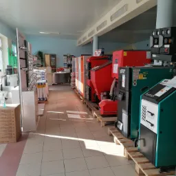 Wnętrze sklepu z ekspozycją różnorodnych modeli kotłów grzewczych na paliwa stałe, ustawionych w rzędzie na drewnianych paletach, widoczne także baterie umywalkowe i wyposażenie łazienkowe.