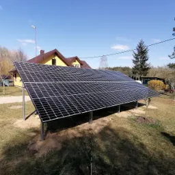 Instalacja fotowoltaiczna na gruncie, panele ustawione pod kątem, widoczny żółty dom jednorodzinny w tle, podłoże piaszczyste wokół słupów wspierających konstrukcję.