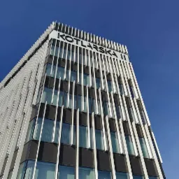 Nowoczesny budynek biurowy z elewacją w odcieniach szarości i czerni, ozdobiony pionowymi, białymi elementami i dużym napisem 'KOTLARSKA' na górze, widziany z dołu na tle błękitnego nieba.