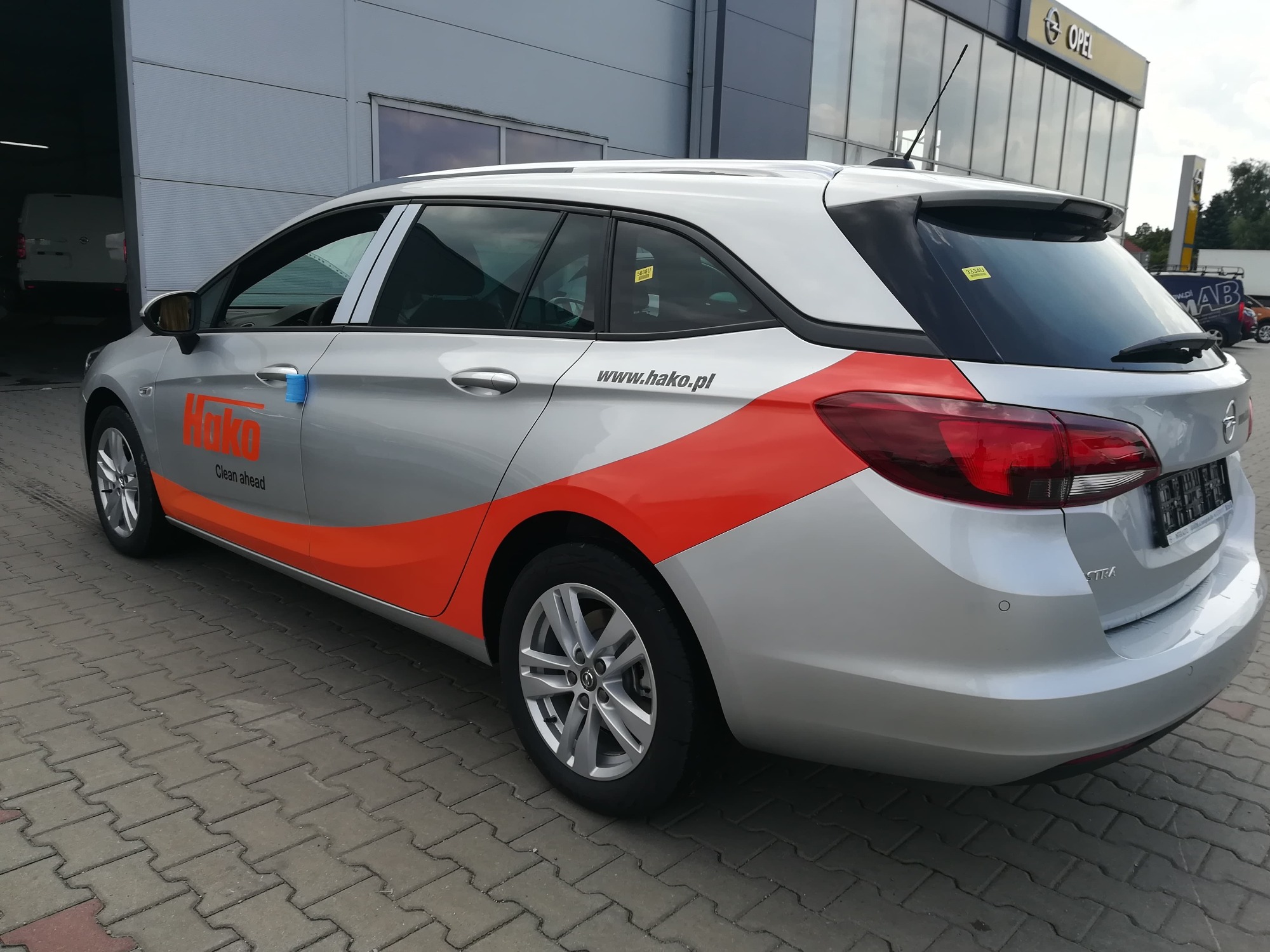Srebrna Opel Astra kombi z pomarańczową grafiką i napisem 'Hako Clean ahead' na boku, zaparkowana przed budynkiem z logo Opel.