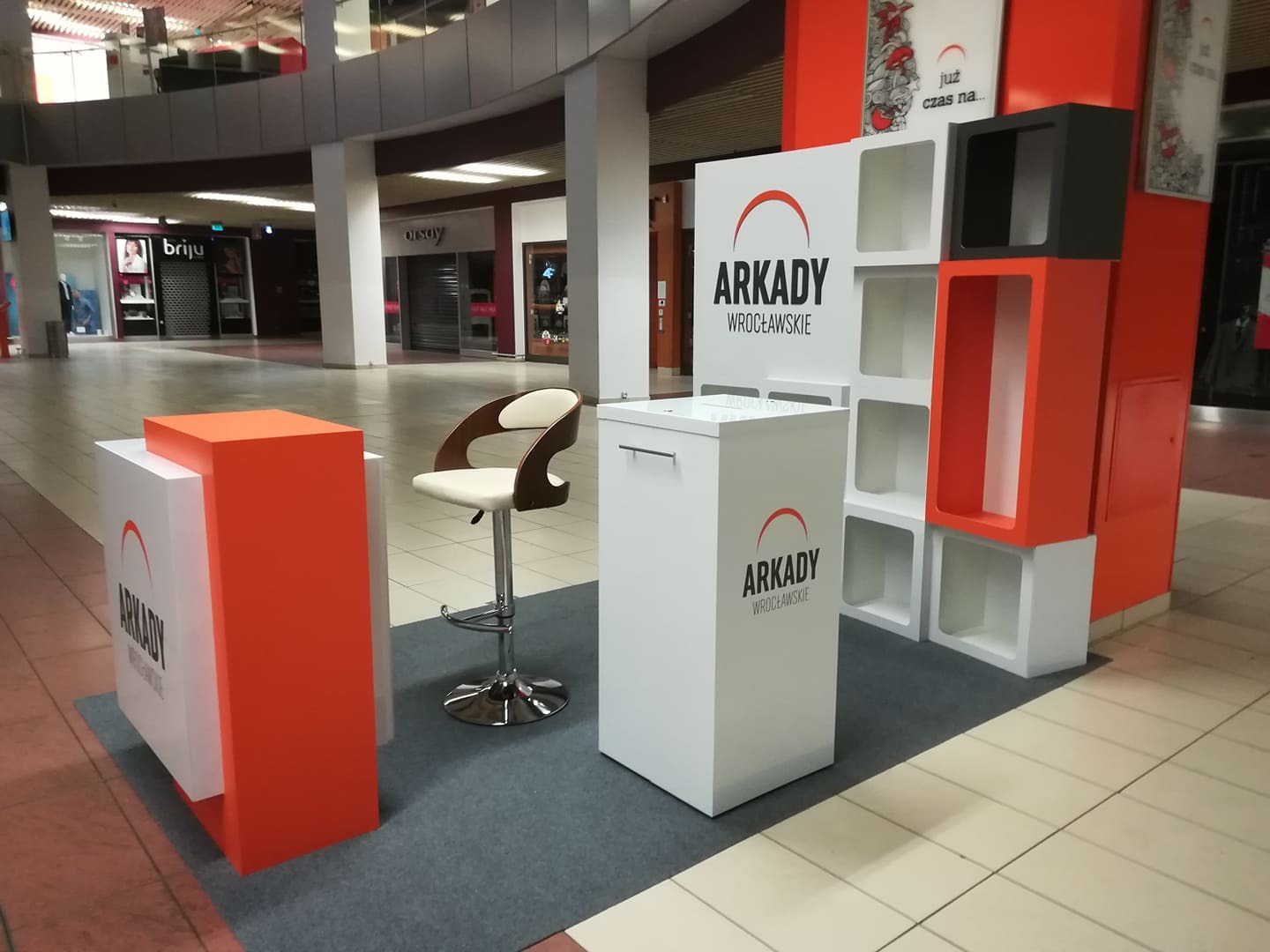 Stoisko reklamowe Arkady Wrocławskie w centrum handlowym, z meblami ekspozycyjnymi w kolorach białym i pomarańczowym, krzesło barowe z brązowymi elementami, widoczne sklepy w tle.