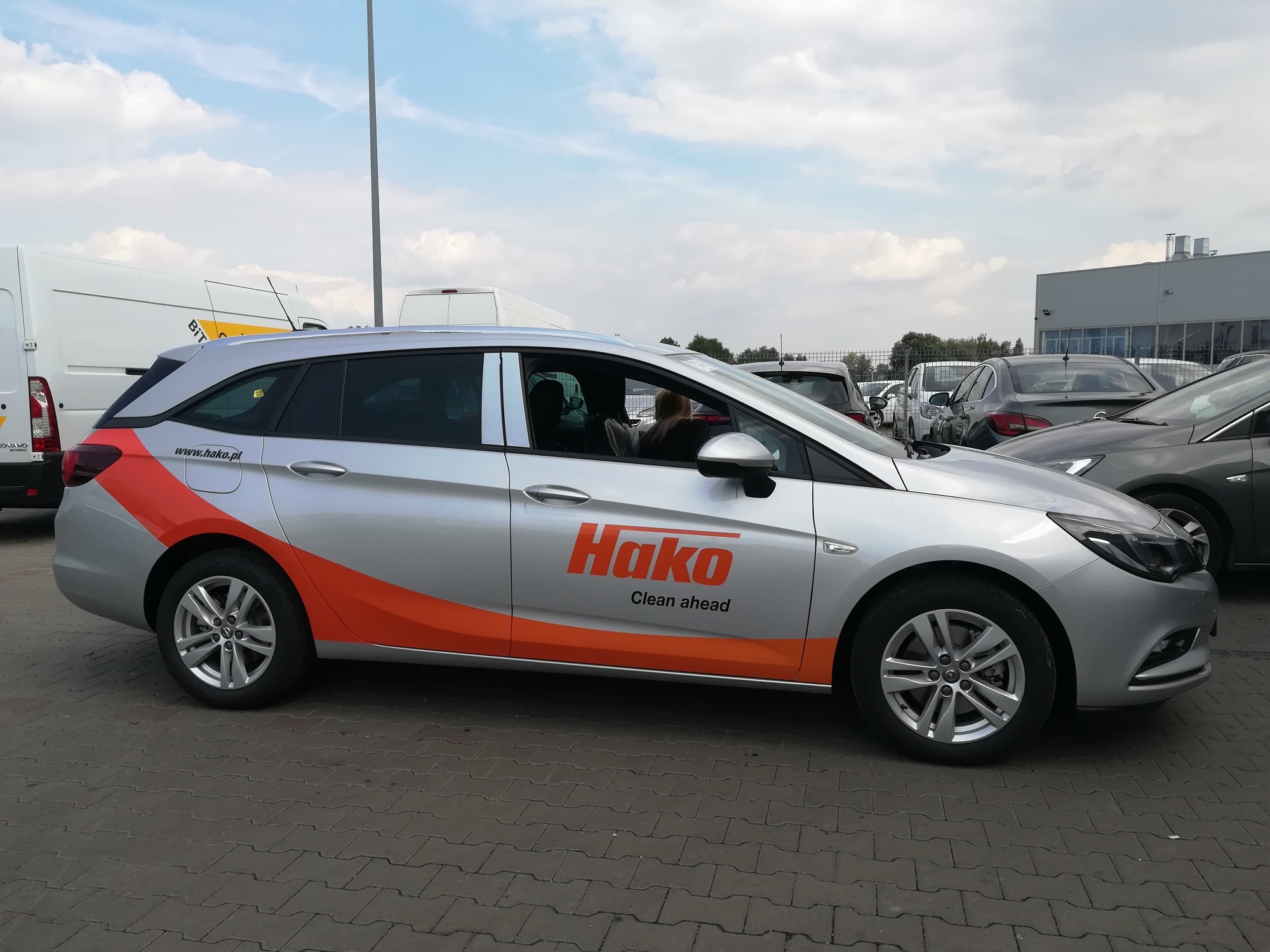 Srebrny samochód typu kombi z pomarańczową grafiką i logo firmy Hako na boku, zaparkowany na brukowanym parkingu z innymi pojazdami w tle.