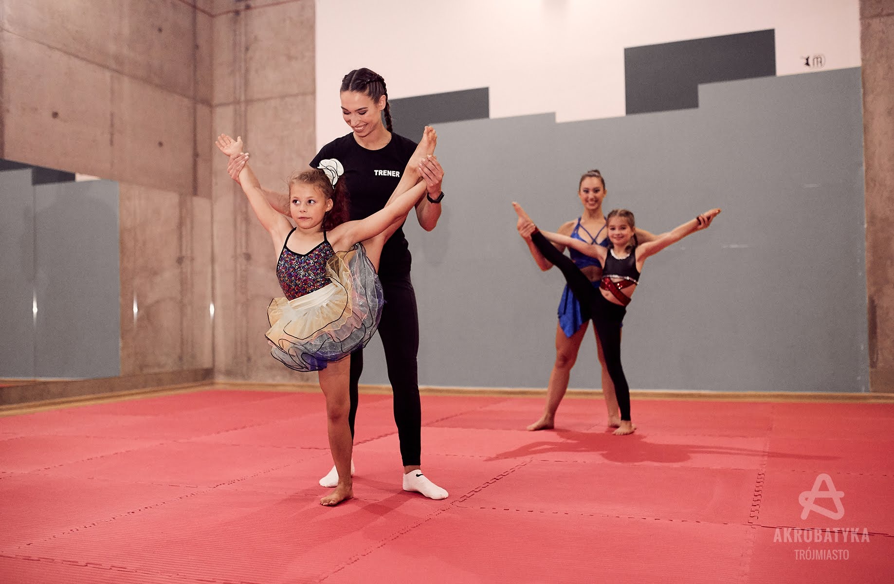Dziewczynka w stroju gimnastycznym ćwiczy rozciąganie z pomocą instruktorki, w tle kolejna para trenująca akrobatykę na czerwonej macie.