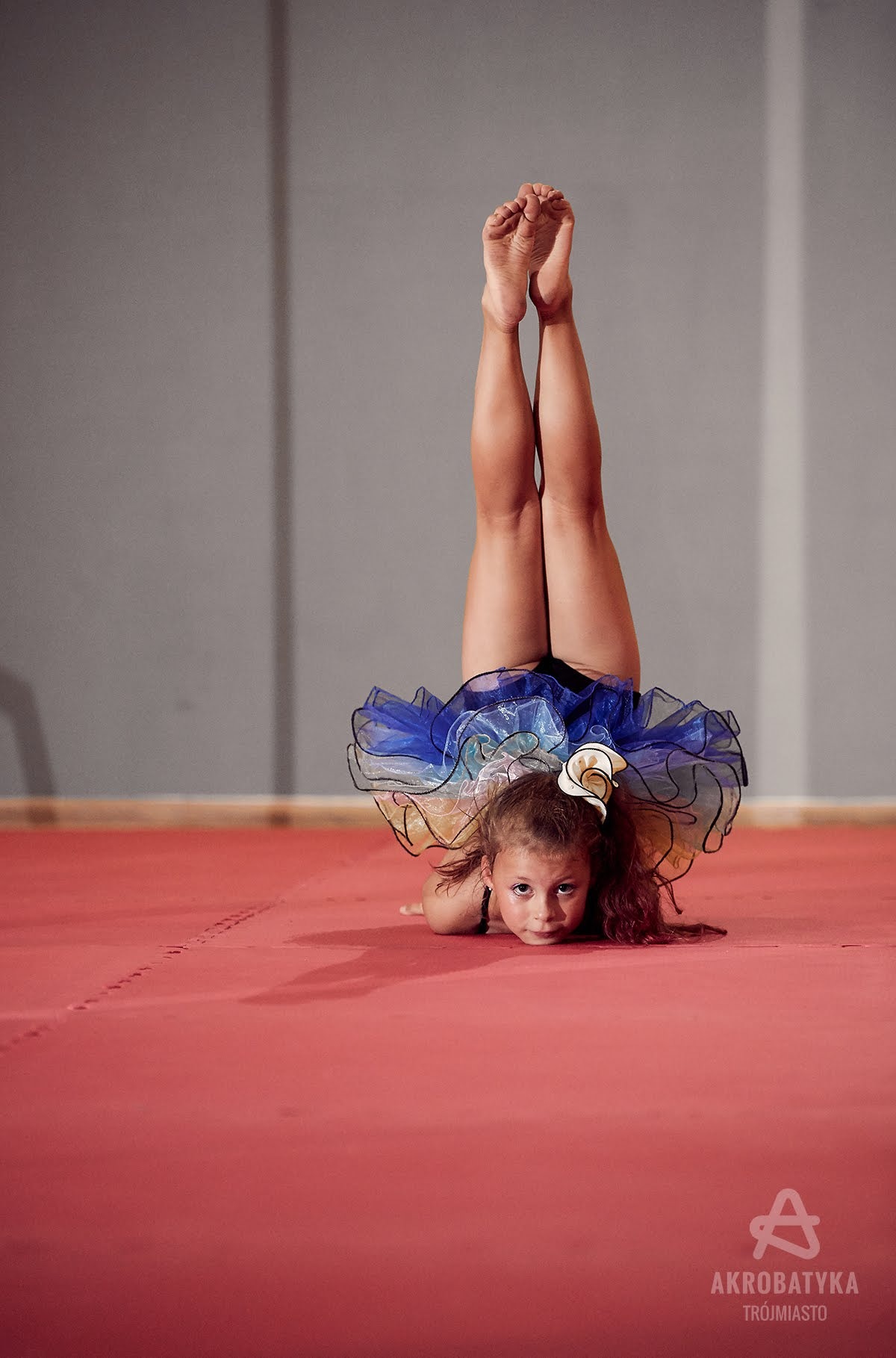 Dziewczynka w stroju gimnastycznym, z nogami w górze, oparta na przedramionach, na czerwonej macie, z logo Akrobatyka Trójmiasto w prawym dolnym rogu.