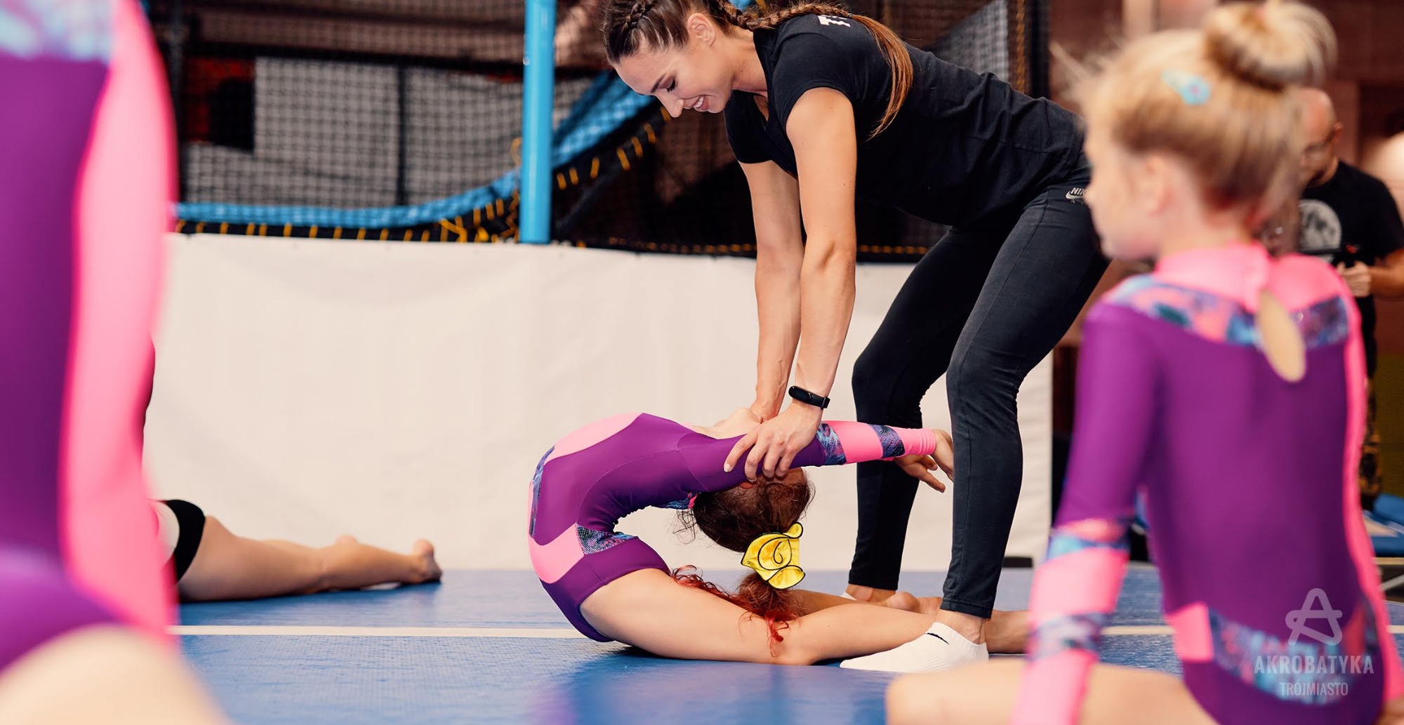 Instruktorka gimnastyki pomaga dziewczynce w fioletowym stroju wykonać mostek na niebieskiej macie w sali gimnastycznej.