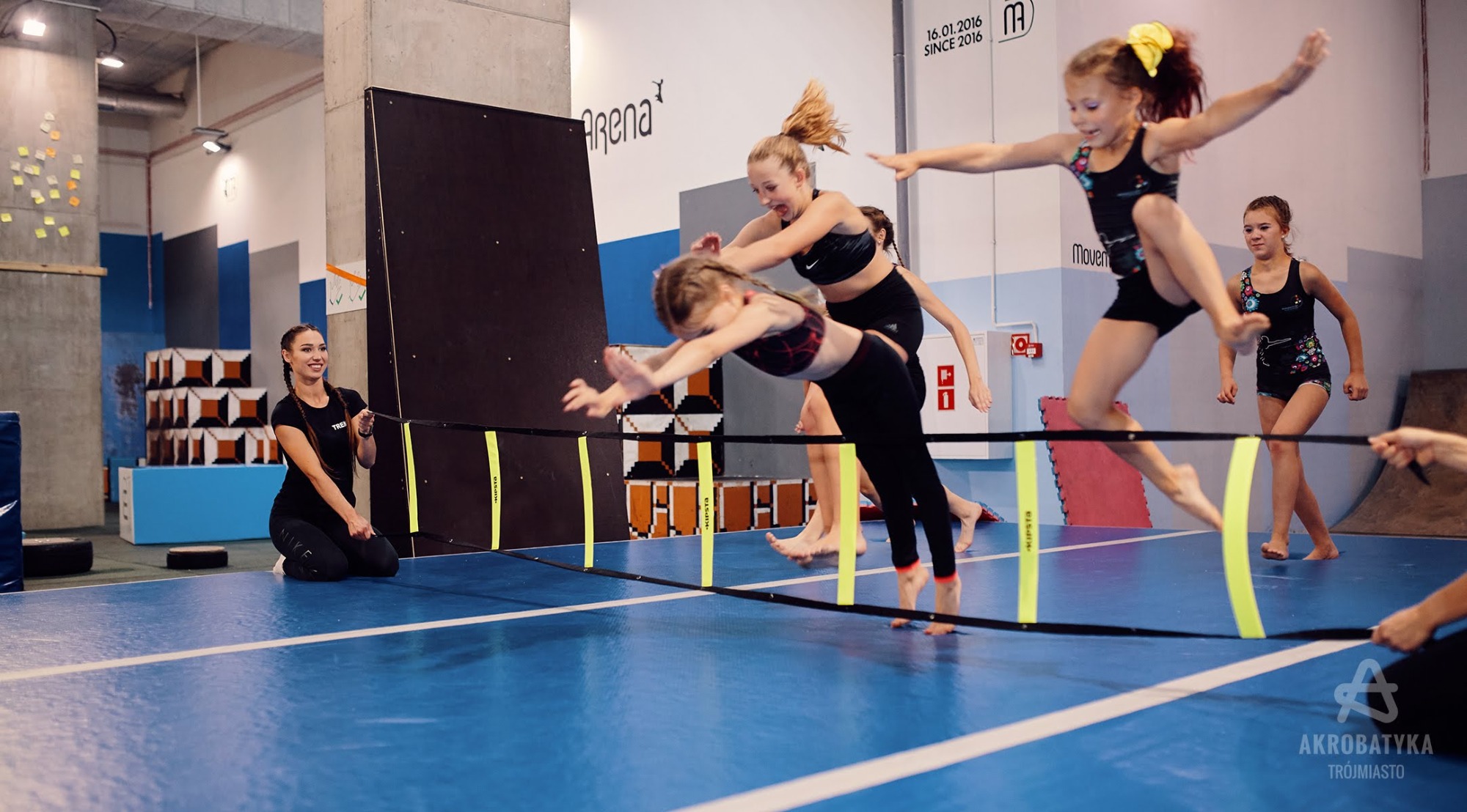 Dziewczęta trenujące akrobatykę skacząc przez przeszkody na niebieskiej macie w hali sportowej, nadzorowane przez instruktorkę.