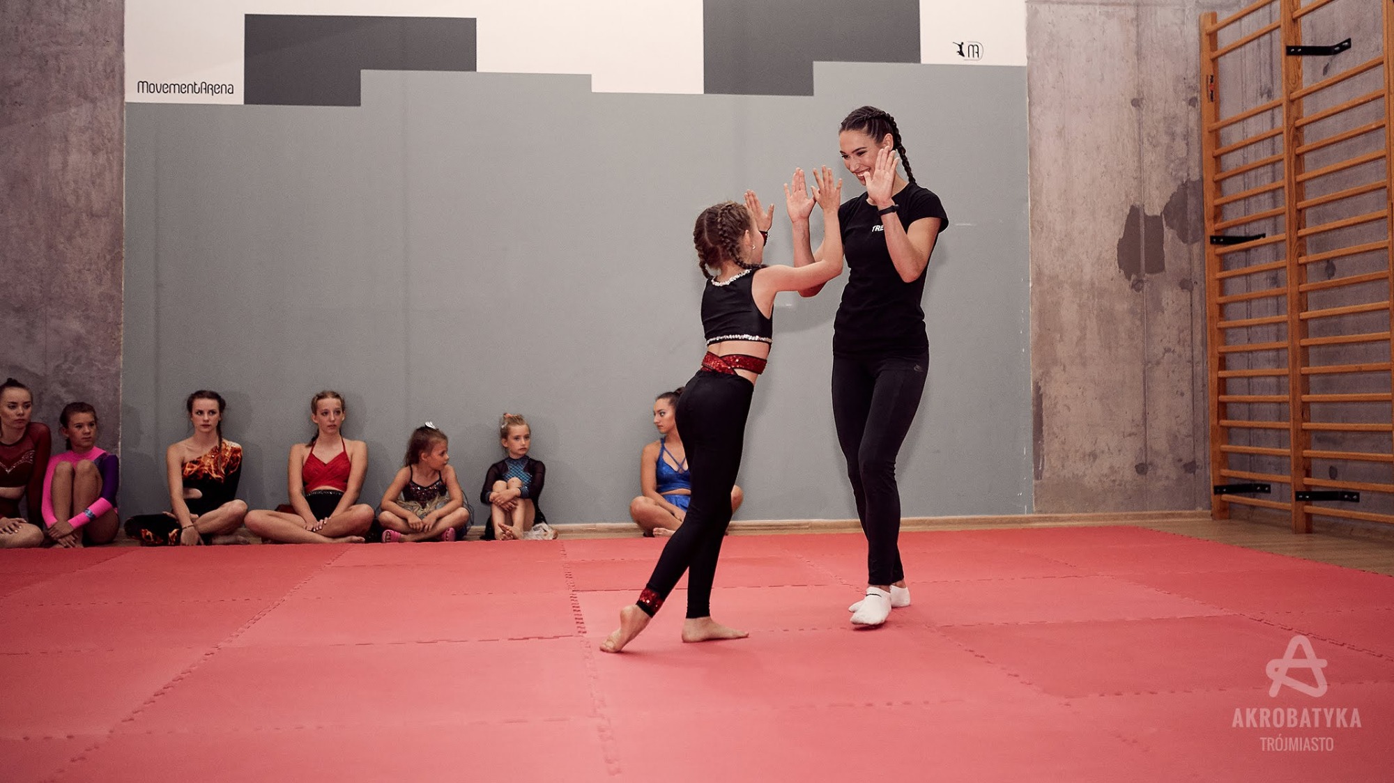 Trenerka personalna z zaplecionymi włosami 'daje piątkę' dziewczynce w sportowym stroju na czerwonej macie w sali gimnastycznej, w tle siedzące dzieci i drabinki gimnastyczne.
