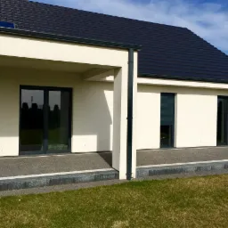 Nowoczesny dom jednorodzinny z białymi ścianami, ciemną dachówką i dużymi przeszkleniami, w tym rolety zewnętrzne częściowo zasłonięte, widok z ogrodu.