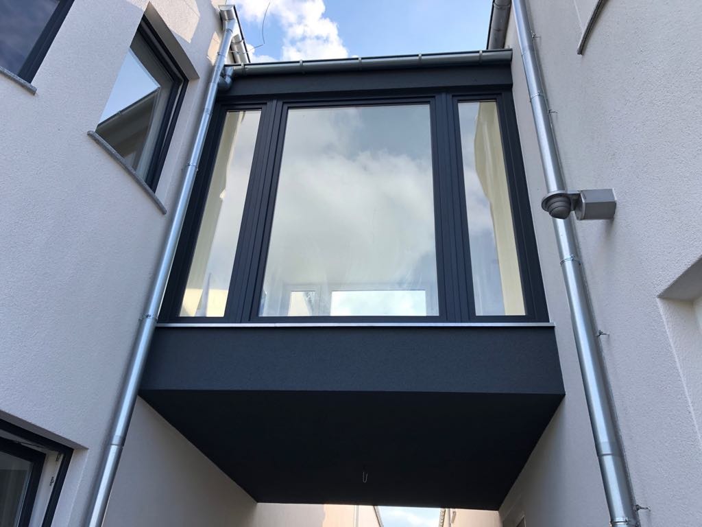 Czarna rama okienna z dużym oknem, montowana pomiędzy dwoma budynkami z białymi ścianami zewnętrznymi, widok na niebo odbijający się w szybie okiennej, rynny odprowadzające wodę po obu stronach.