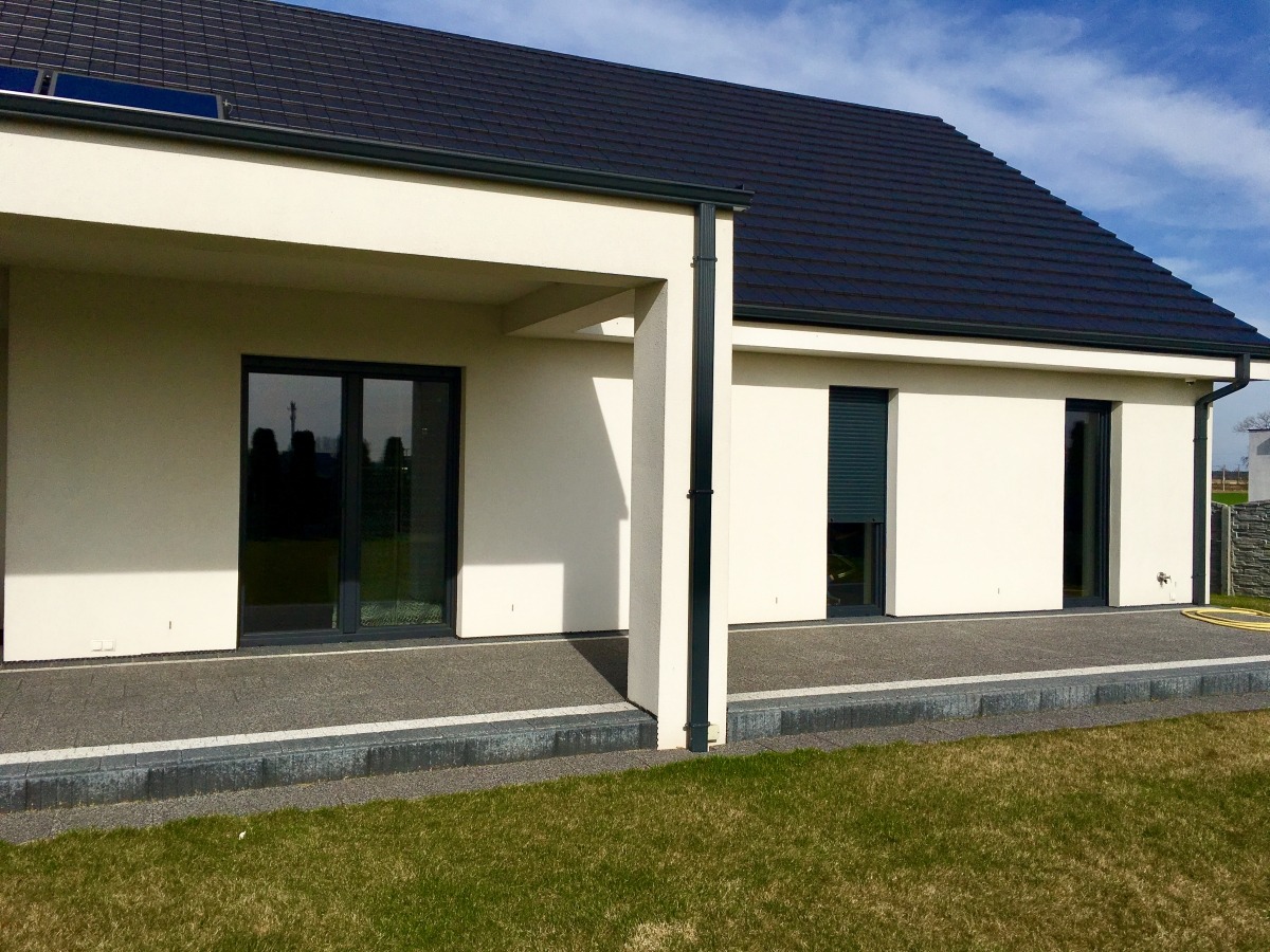 Nowoczesny dom jednorodzinny z białymi ścianami, ciemną dachówką i dużymi przeszkleniami, w tym rolety zewnętrzne częściowo zasłonięte, widok z ogrodu.