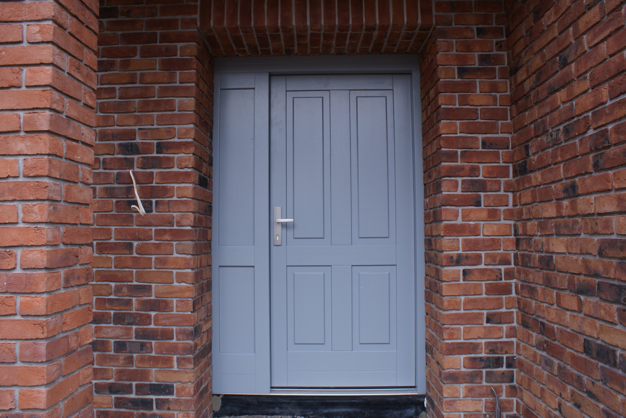 Szare drzwi wejściowe z panelem bocznym, osadzone w murze z czerwonej cegły, z widocznym fragmentem przewodu elektrycznego wystającego ze ściany.