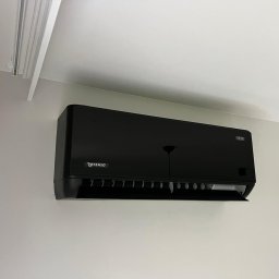 Klimatyka.com Bartłomiej Szrejner - Czarna jednostka klimatyzacji Rotenso TETA zamontowana na jasnej ścianie w pomieszczeniu. Widoczne żaluzje w dolnej części urządzenia.