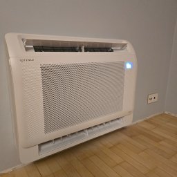 Klimatyka.com Bartłomiej Szrejner - Biała, naścienna jednostka klimatyzacyjna ROTENSG z niebieską diodą, zainstalowana w pomieszczeniu z drewnianą podłogą i szarą ścianą.