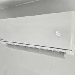 Biała jednostka wewnętrzna klimatyzatora Midea, zamontowana na szarej ścianie, wyświetlająca temperaturę 16 stopni Celsjusza.