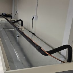 Klimatyka.com Bartłomiej Szrejner - Widok z góry na fragment instalacji wentylacyjnej z miedzianymi rurami w czarnej izolacji, zamontowanej pod sufitem podwieszanym w jasnym pomieszczeniu biurowym.