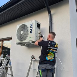 Klimatyka.com Bartłomiej Szrejner - Instalator na drabinie montuje zewnętrzną jednostkę klimatyzacji AUX na elewacji budynku. Widoczne narzędzia i elementy instalacji. Ujęcie z dołu, błękitne niebo w tle.