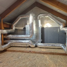Klimatyka.com Bartłomiej Szrejner - Kompletna instalacja rekuperacji na poddaszu z widocznymi przewodami w srebrnej izolacji i centralą rekuperacyjną. Drewniana konstrukcja dachu w tle.