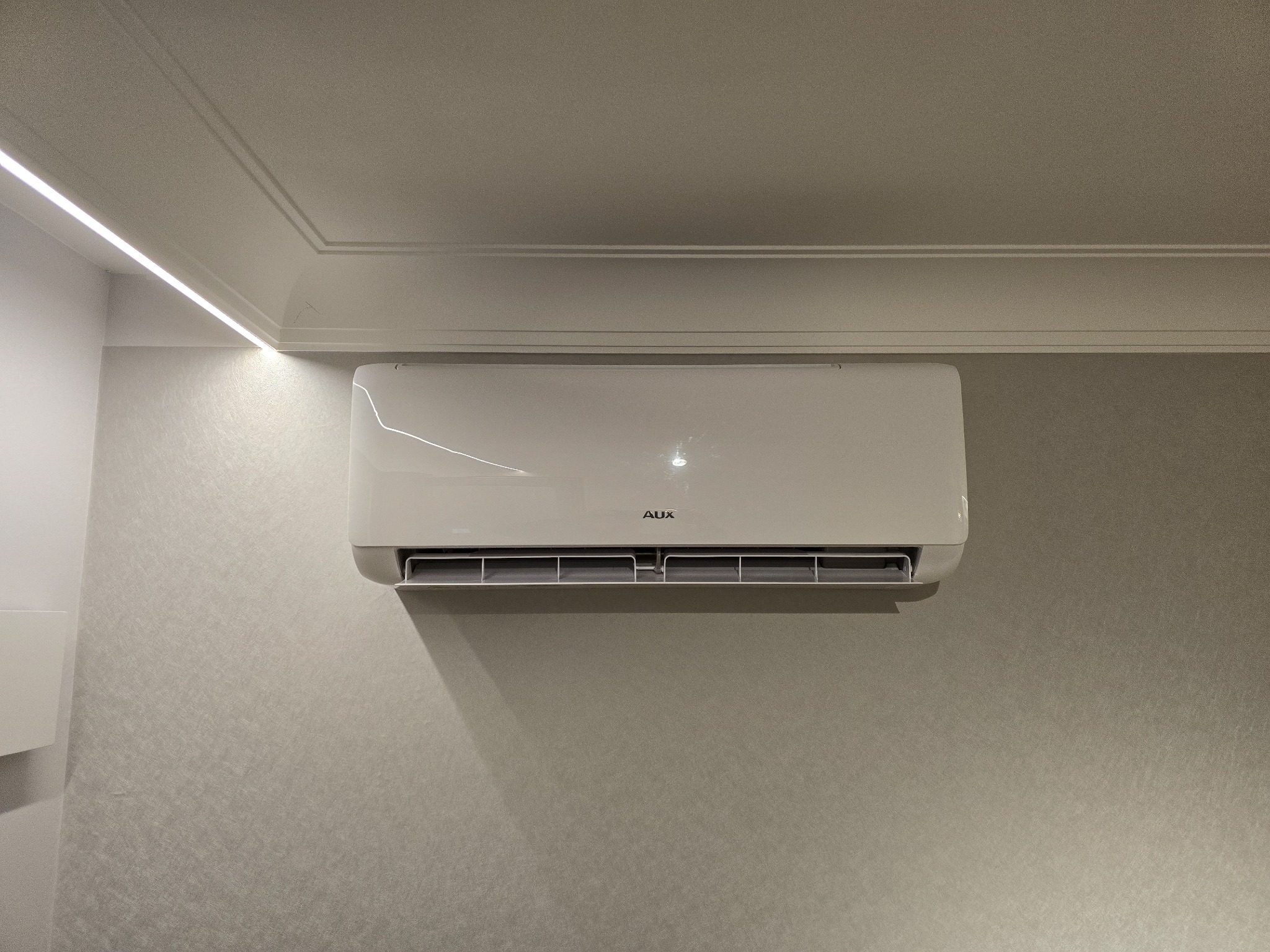 Biały klimatyzator AUX zamontowany na ścianie z delikatną teksturą, oświetlony listwą LED pod sufitem z dekoracyjnym gzymsem.