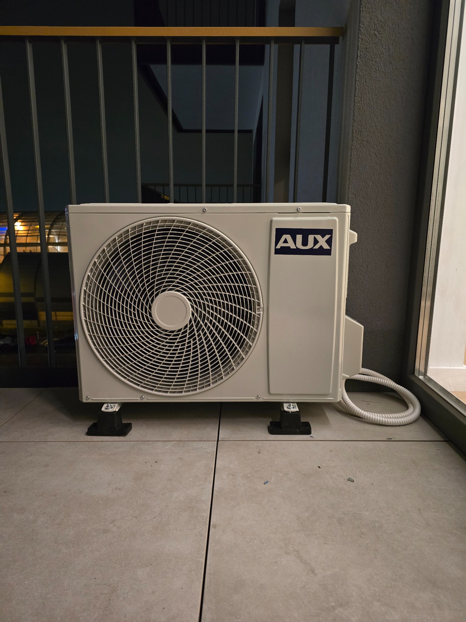 Biała jednostka zewnętrzna klimatyzatora marki AUX zamontowana na balkonie, widoczne żaluzje balkonu i fragment okna.