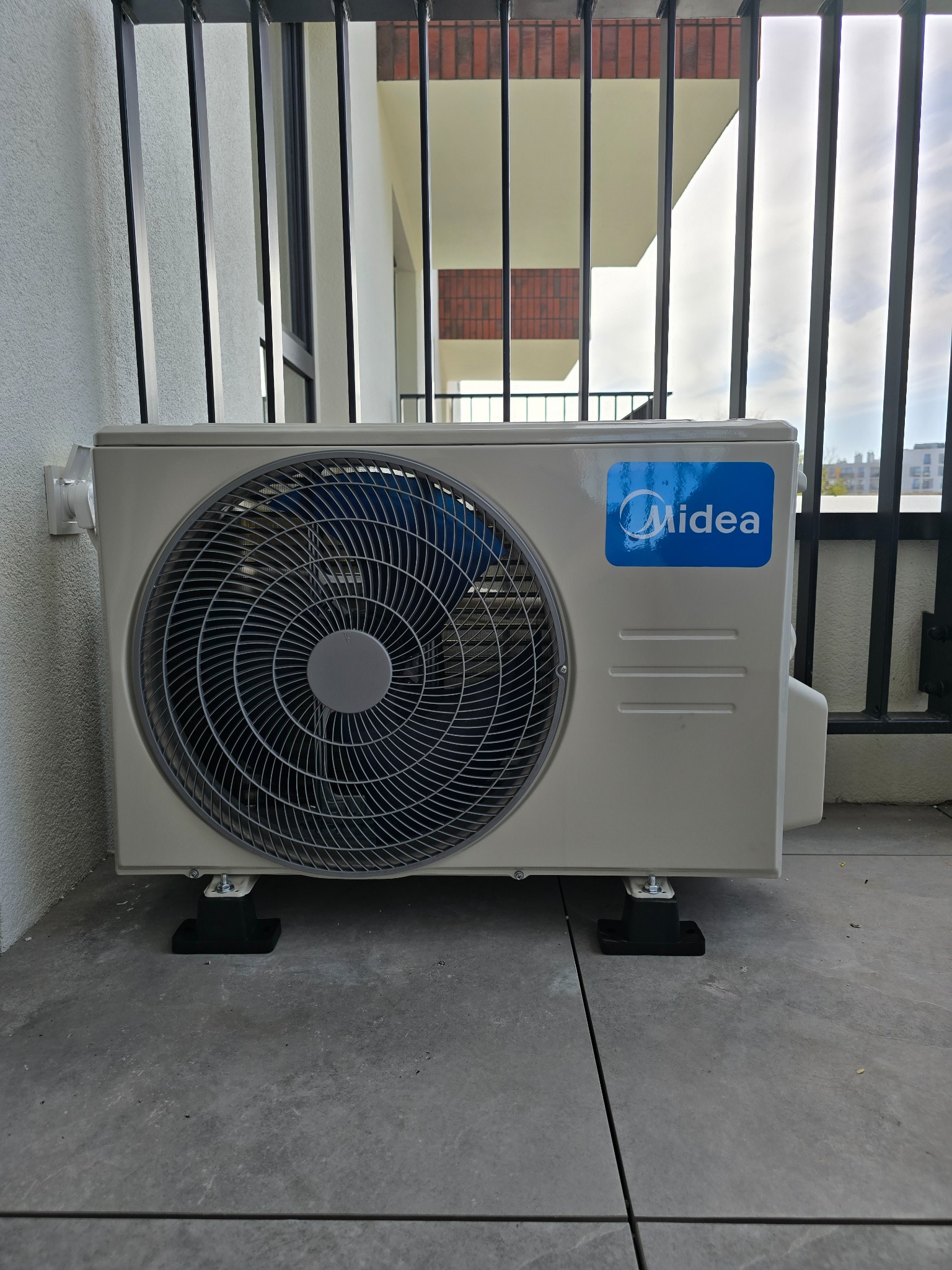 Zewnętrzna jednostka klimatyzacji Midea zamontowana na balkonie, widoczne czarne gumowe nóżki antywibracyjne i metalowa balustrada.