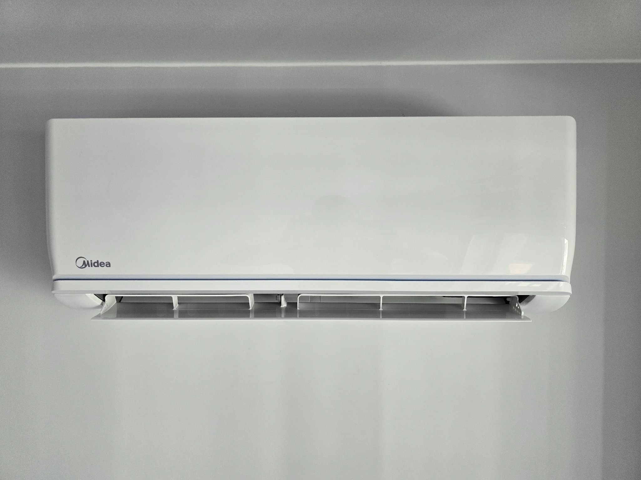 Biała jednostka klimatyzacji Midea zamontowana na szarej ścianie, widoczne otwarte żaluzje wylotu powietrza.