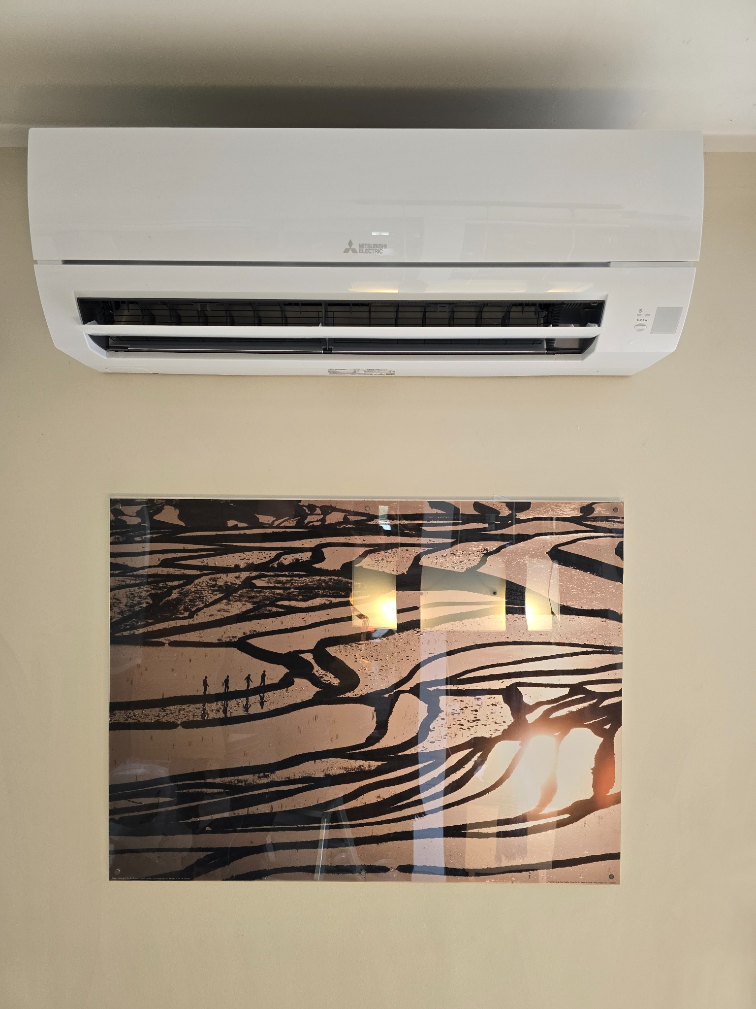 Biały klimatyzator Mitsubishi Electric zamontowany na beżowej ścianie nad obrazem przedstawiającym krajobraz z ludźmi.