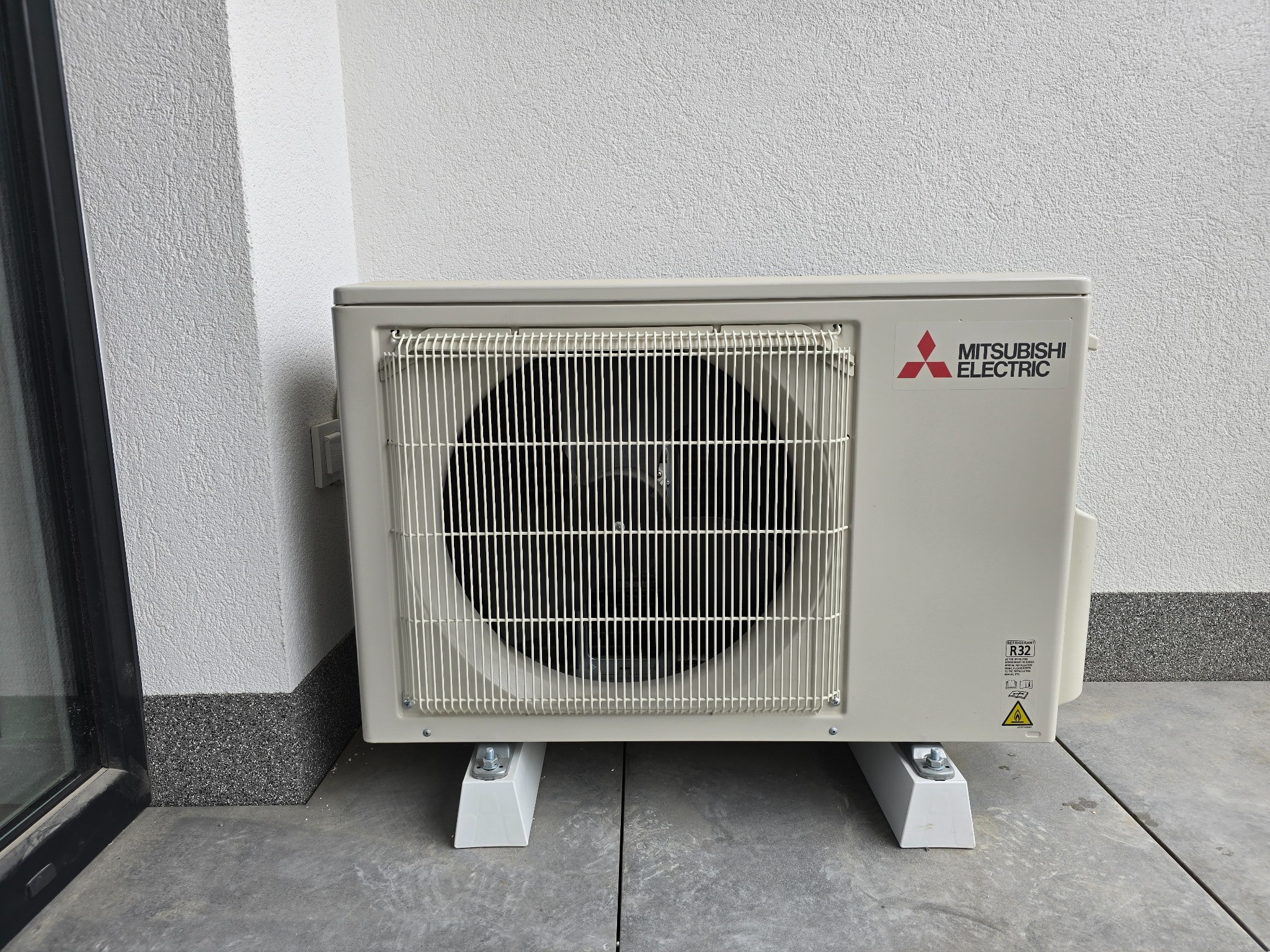 Zewnętrzna jednostka klimatyzacji Mitsubishi Electric, zamontowana na białych wspornikach na szarym betonowym podłożu, przy ścianie z szarą, teksturowaną listwą przypodłogową.