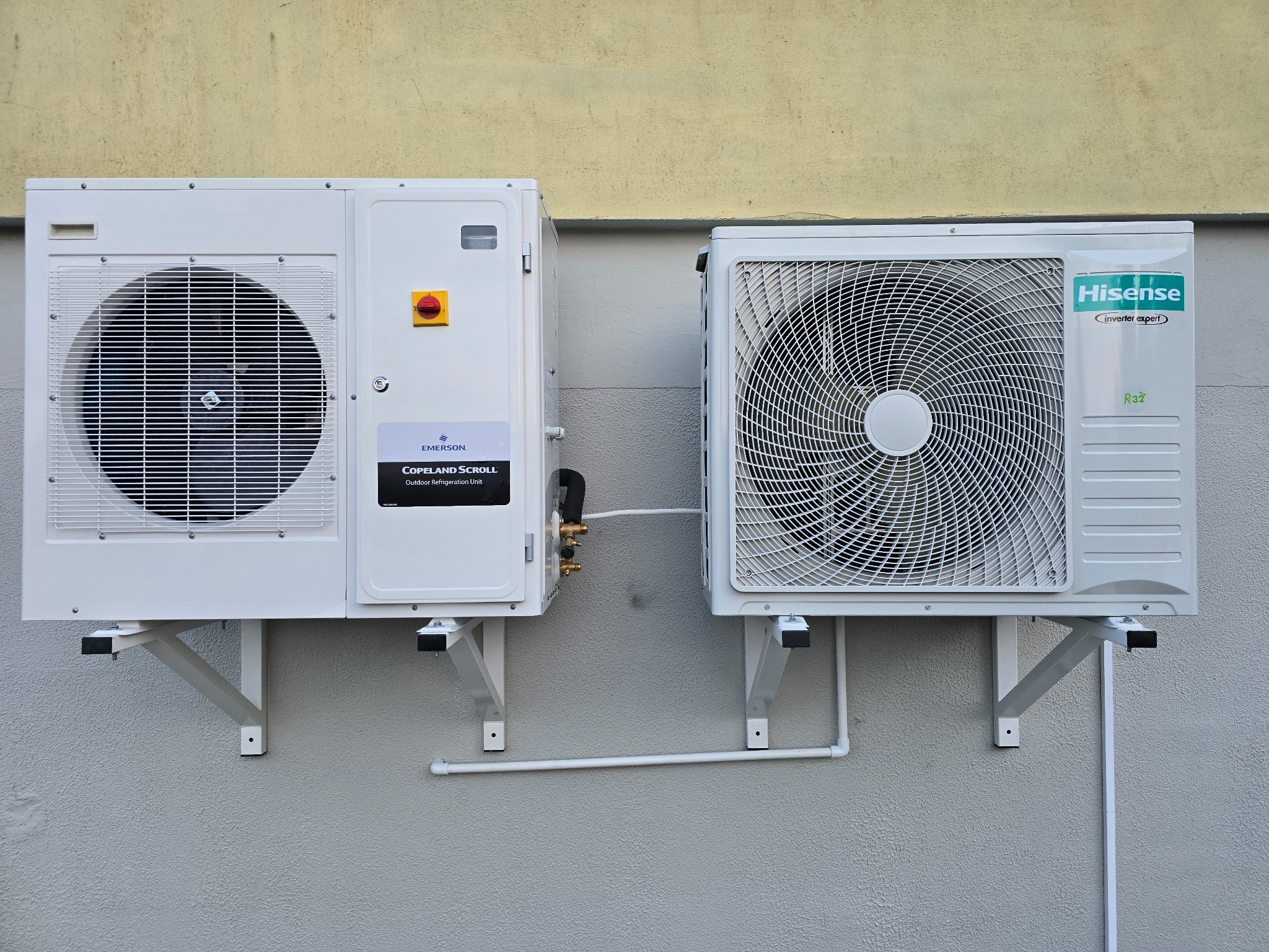 Dwa białe agregaty klimatyzacyjne Hisense i Emerson zamontowane na wspornikach na szarej ścianie zewnętrznej budynku, widoczne przewody chłodnicze i instalacja odprowadzająca skropliny.