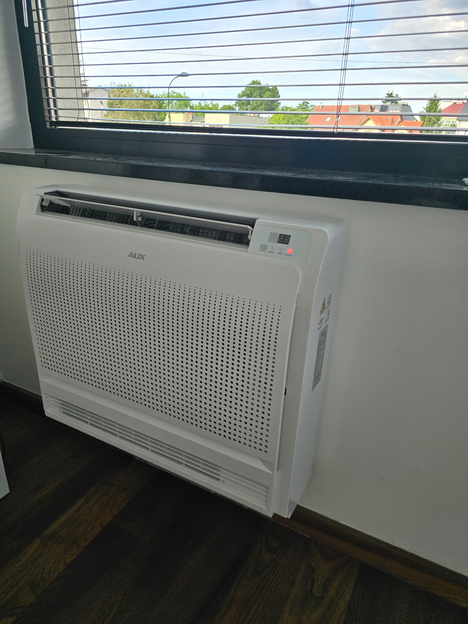 Biały klimatyzator podłogowy marki AUX zamontowany przy ścianie, widoczny wyświetlacz z czerwoną diodą, w tle okno z żaluzjami i widok na zabudowania.