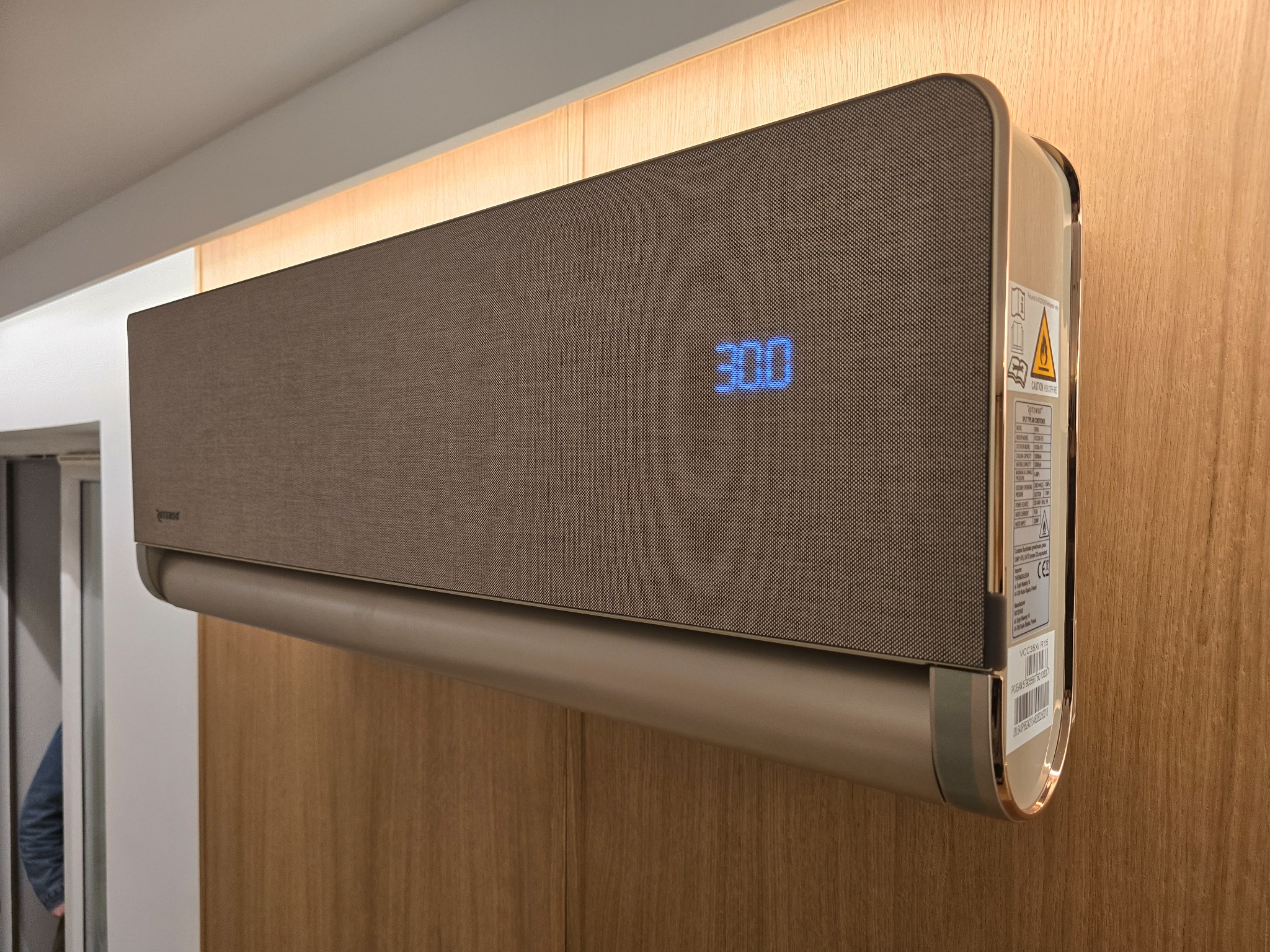 Nowoczesny klimatyzator ścienny w kolorze beżowym z wyświetlaczem temperatury 30.0 stopni Celsjusza, zamontowany na drewnianej ścianie z subtelnym oświetleniem LED w tle.