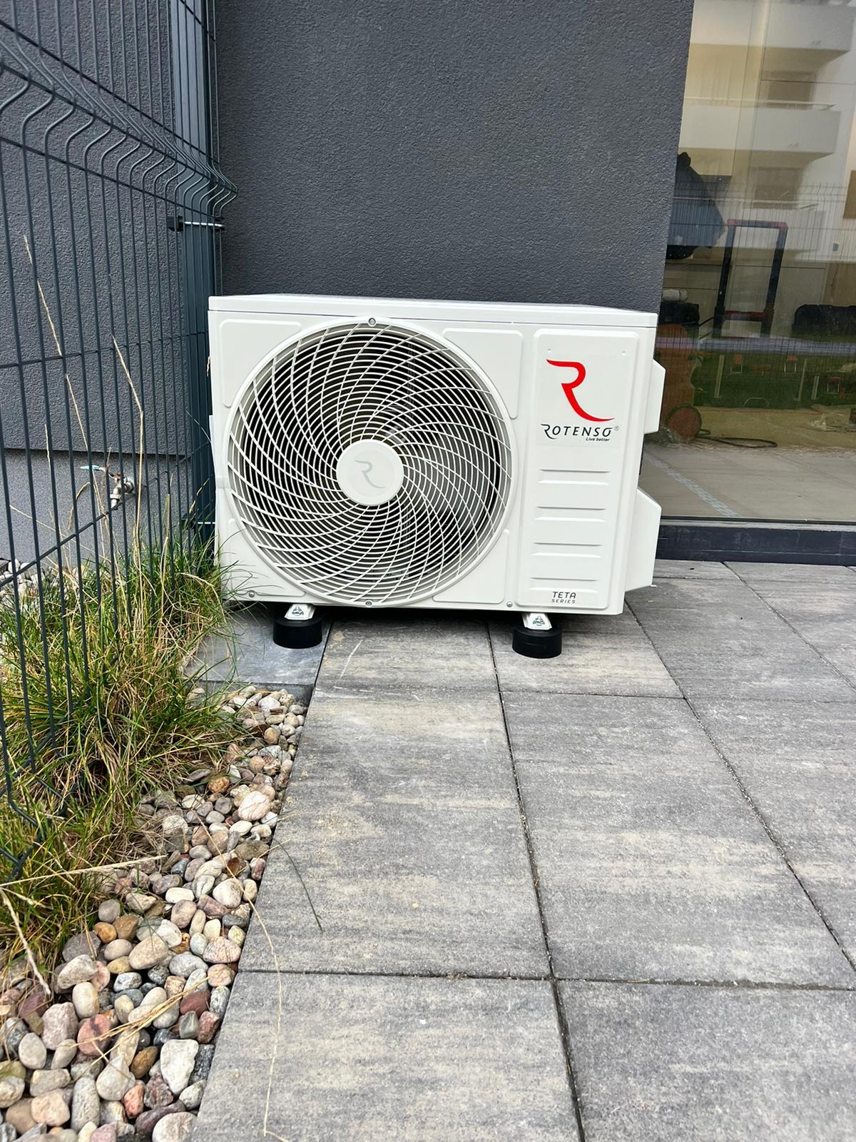 Biała jednostka zewnętrzna klimatyzatora marki Rotenso, model Teta, umieszczona na czarnych podkładkach antywibracyjnych, stoi na szarych płytach chodnikowych obok ogrodzenia panelowego i ściany...