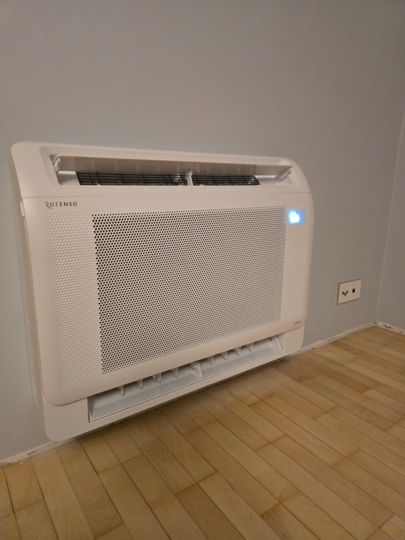 Biała, naścienna jednostka klimatyzacyjna ROTENSG z niebieską diodą, zainstalowana w pomieszczeniu z drewnianą podłogą i szarą ścianą.