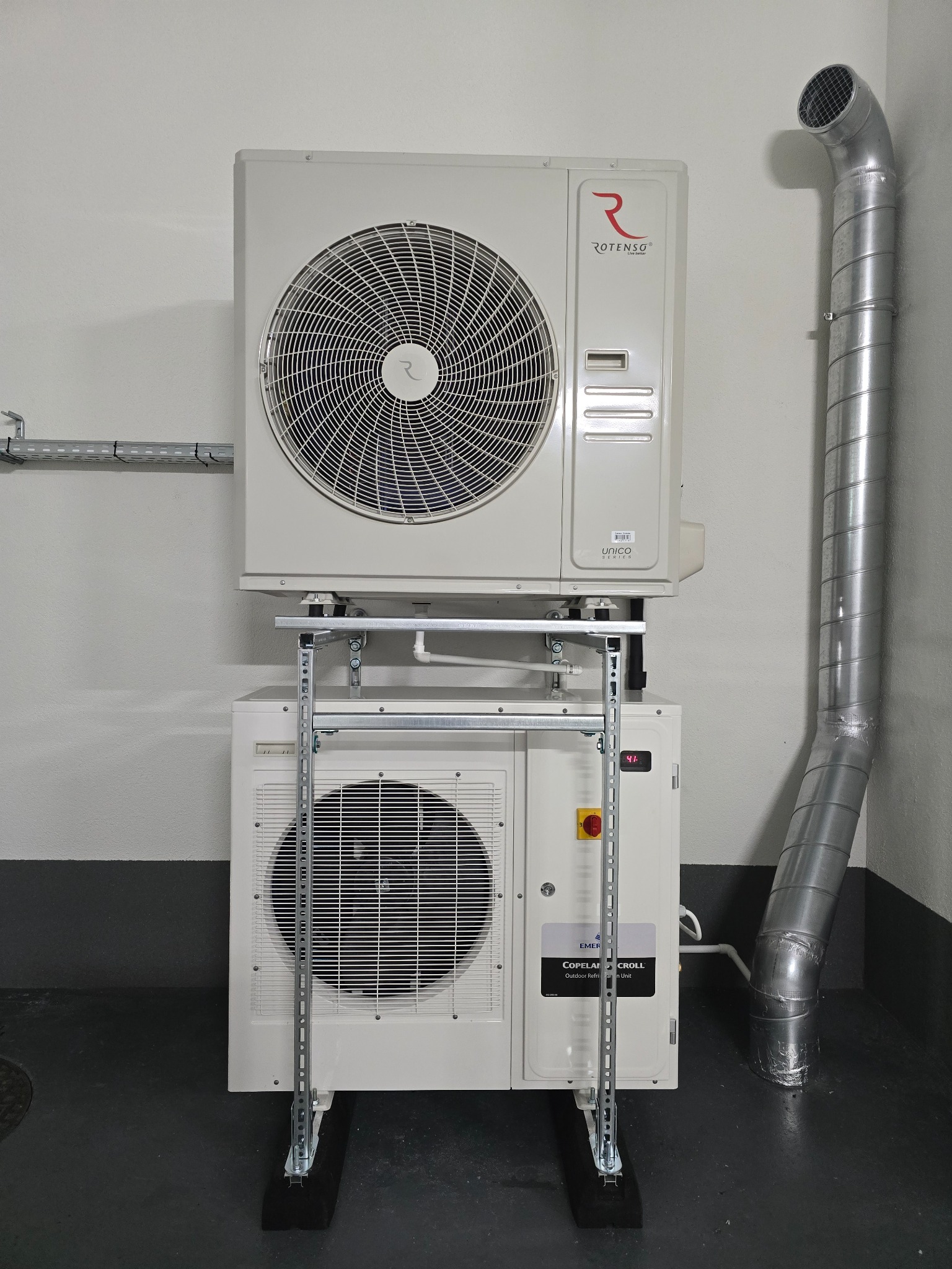 Dwa białe urządzenia klimatyzacyjne marki Rotenso i Copeland Scroll, zamontowane jedno nad drugim na metalowym stelażu w pomieszczeniu technicznym, z widoczną rurą wentylacyjną.