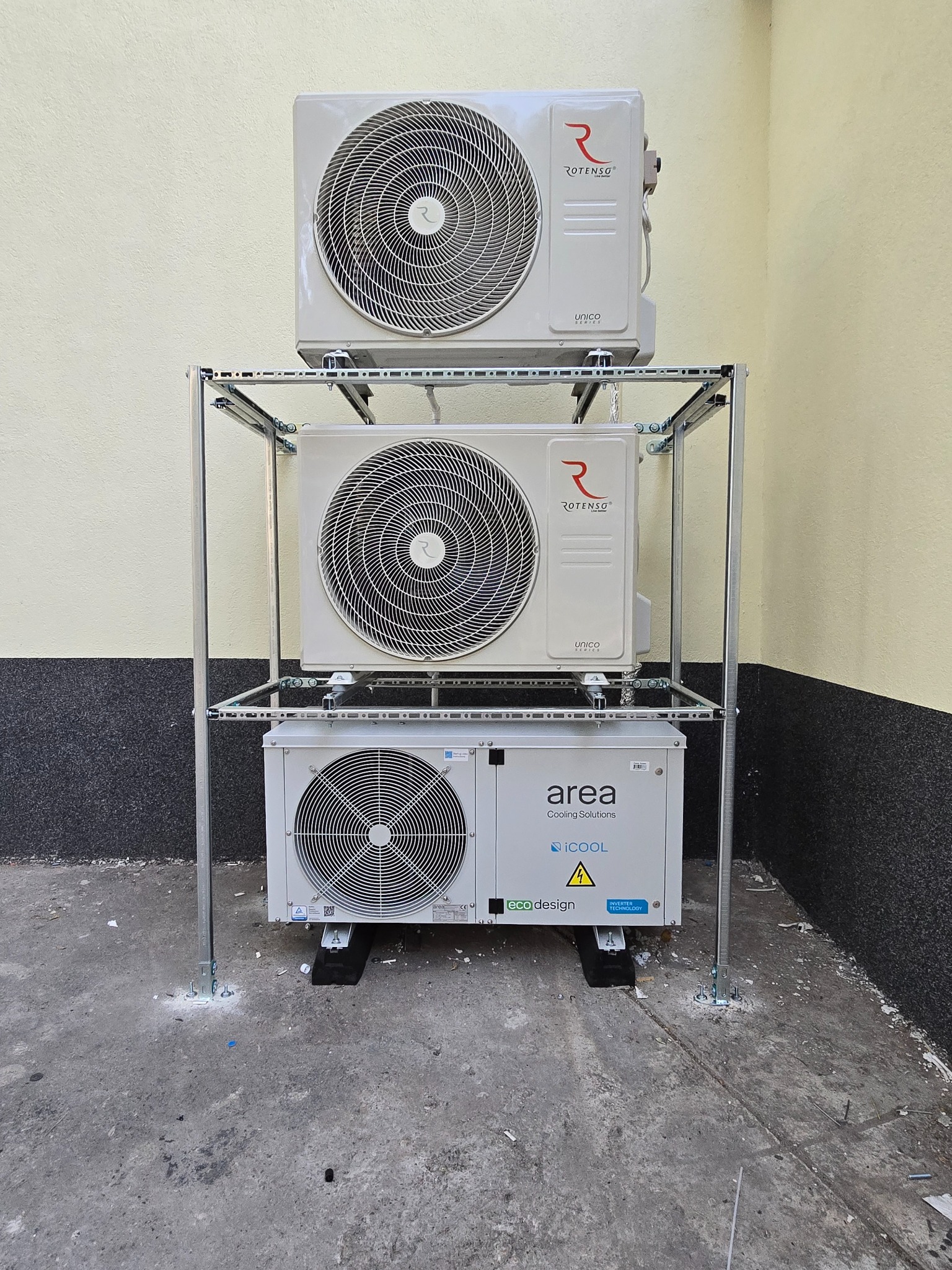 Trzy jednostki zewnętrzne klimatyzacji zamontowane jedna nad drugą na metalowej konstrukcji wsporczej przy ścianie budynku. Dwie górne jednostki marki Rotenso, dolna marki Area.