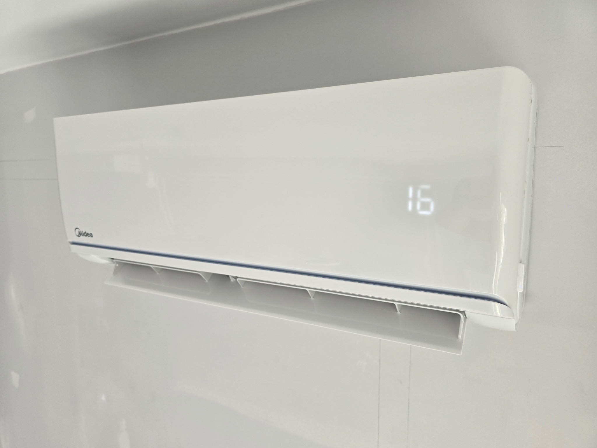 Biała jednostka wewnętrzna klimatyzatora Midea, zamontowana na szarej ścianie, wyświetlająca temperaturę 16 stopni Celsjusza.