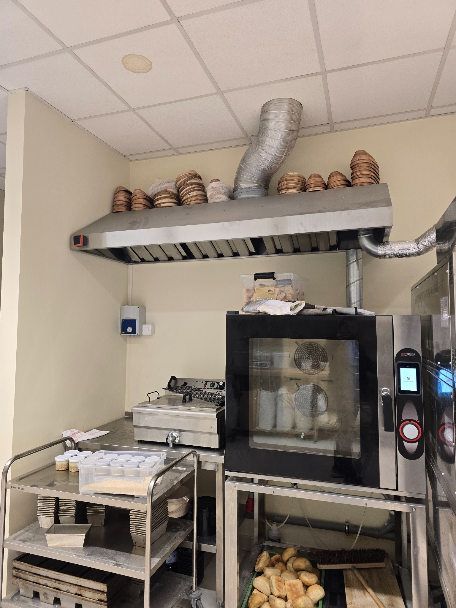 Profesjonalna kuchnia z nierdzewną stalą: okap wentylacyjny z rurą, piec konwekcyjny z widocznymi wentylatorami i workami w środku, oraz wózki z pojemnikami i formami do pieczenia, a także kosz...