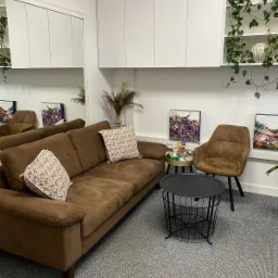 Poczekalnia z brązową sofą, wzorzystymi poduszkami, okrągłym stolikiem kawowym z czarnego metalu, brązowym krzesłem i dekoracjami ściennymi w jasnym pomieszczeniu.