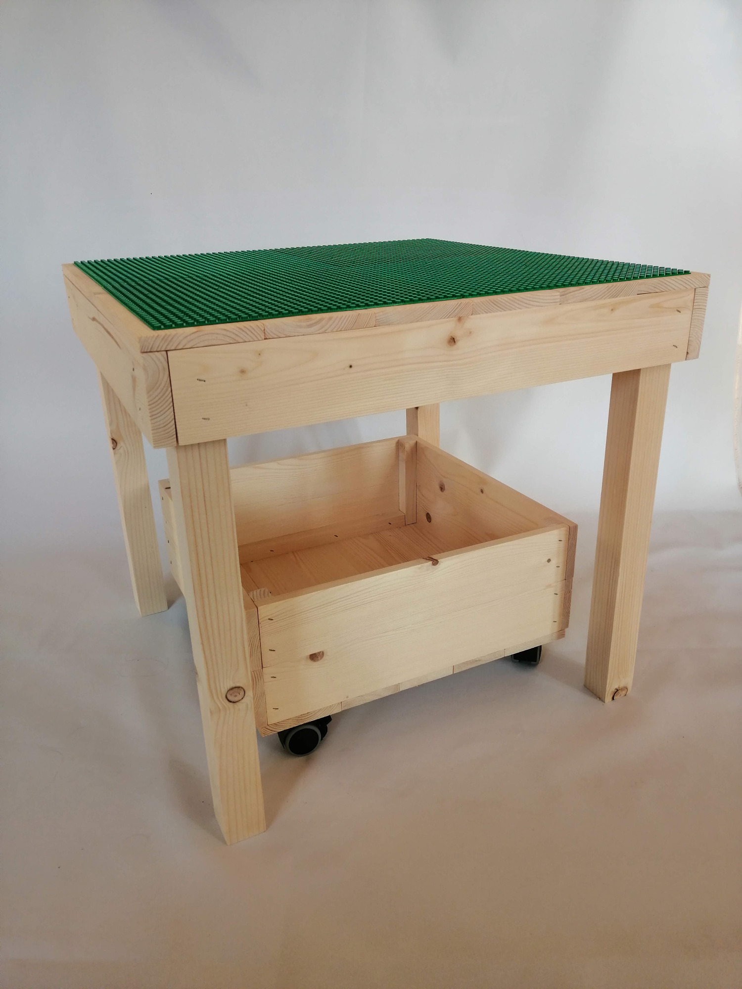 Surowy drewniany stolik dla dzieci z zieloną płytą kompatybilną z klockami, wyposażony w mobilny pojemnik na klocki na kółkach.