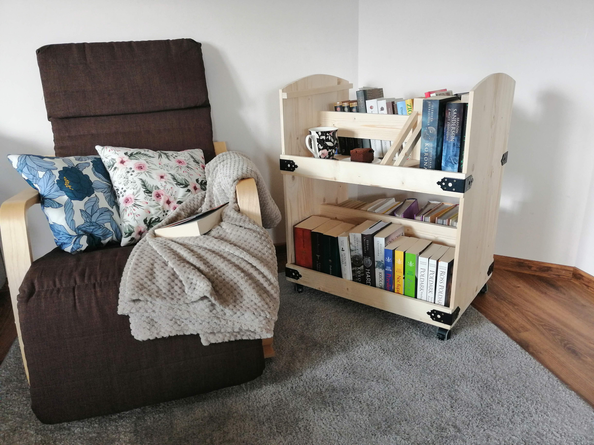 Drewniana, dwupoziomowa biblioteczka mobilna na kółkach, wypełniona książkami i drobnymi przedmiotami, stoi obok fotela z poduszkami w kwiatowe wzory i pledem. Biblioteczka wykończona czarnymi...